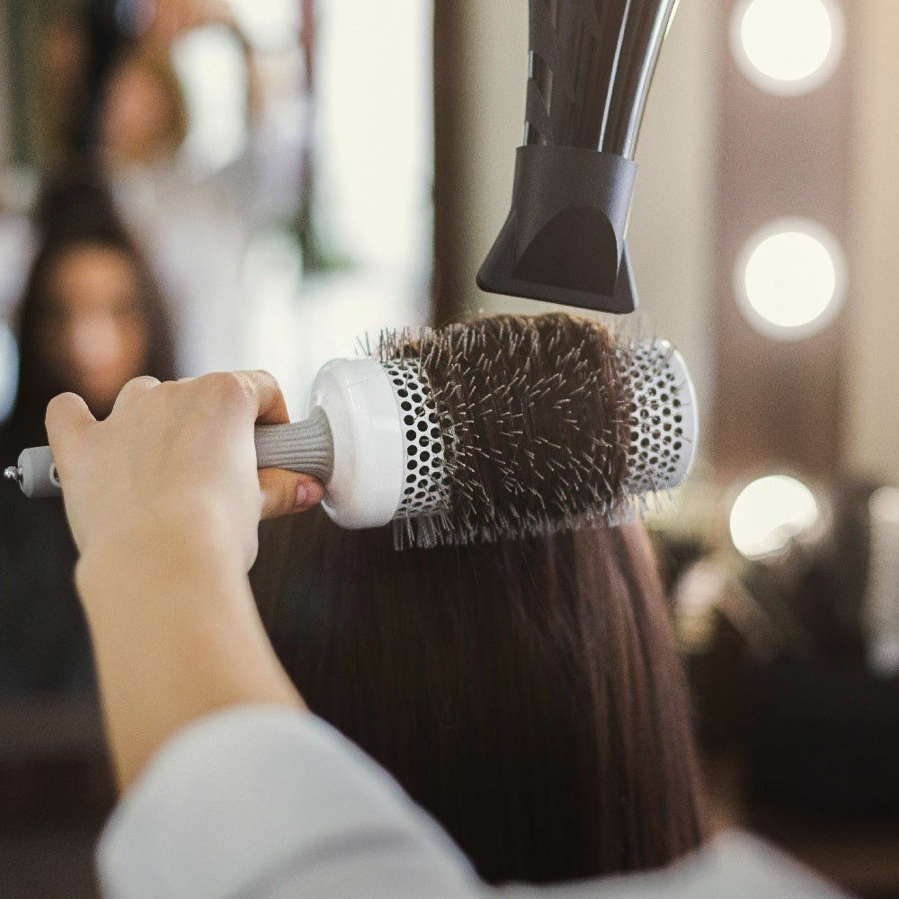 Lady holding a blow drying brush with her left hand and dryer with her right hand to style a client's neck length hair by a vanity station. 